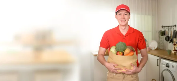 Portrait of Asian young man delivery in red uniform holding healthy fruit vegetables in grocery bag over kitchen background