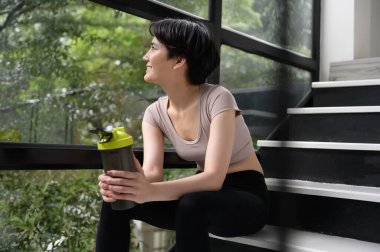 Asyalı genç bir kadın protein takviyesi içiyor, evde egzersiz yapıyor, sağlıklı yaşam tarzı kavramı.