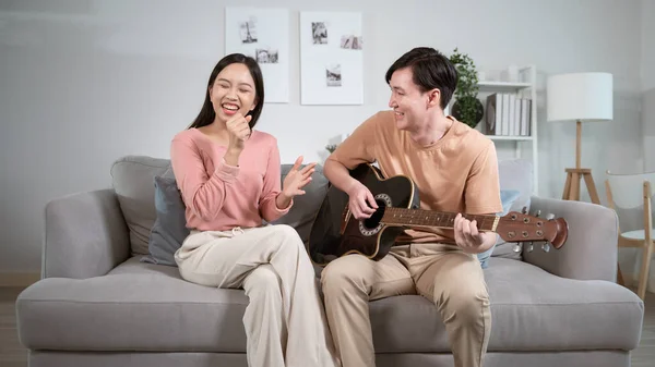 Happy young Asian couple singing a song playing  guitar on sofa in living room at hom