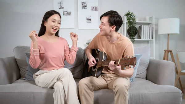 Happy young Asian couple singing a song playing  guitar on sofa in living room at hom