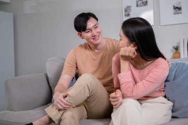 Happy young Asian couple sitting on sofa relaxing together in living room at home