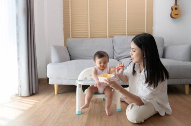 Genç bir anne bebek koltuğunda bebek maması yemesine yardım ediyor.