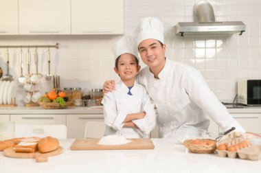 Genç Asyalı baba ve oğlu aşçı üniforması giyip evde birlikte yemek pişiriyorlar.