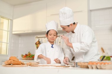 Genç Asyalı baba ve oğlu aşçı üniforması giyip evde birlikte yemek pişiriyorlar.