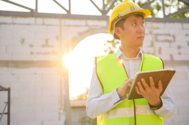 Asyalı bir mühendis kafasına koruyucu bir kask takıyor. Tablet Analitik Mühendisliği verilerini kullanıyor.