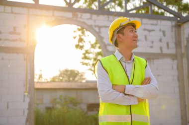 Güvenlik kaskı takan Asyalı bir mühendisin portresi inşaat alanını kontrol ediyor ve proje ilerlemesini analiz ediyor. 
