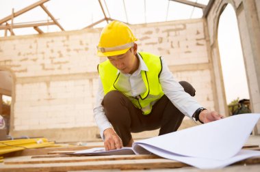 Güvenlik kaskı takan Asyalı bir mühendis inşaat alanındaki çizimleri analiz ediyor.