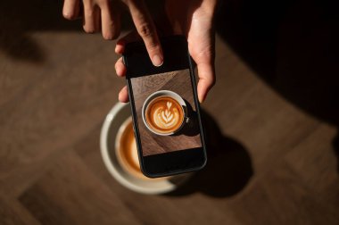 Akıllı telefon, ahşap arka planda latte resimli sıcak kapuçino çekiyor.