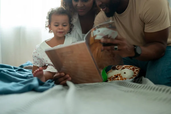 Happy African American Parents with  little daughters playing , reading book on the bed in bedroom at home, happy family concept
