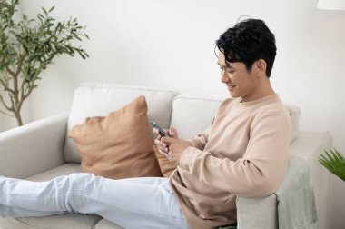 Mutlu Asyalı bir adam evdeki oturma odasında akıllı telefon kullanarak kanepede oturuyor, rahatlıyor zaman ve yaşam tarzı kavramı