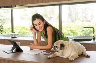 Genç Asyalı kadın tabletle çalışıyor ve evde köpeğiyle eğleniyor.