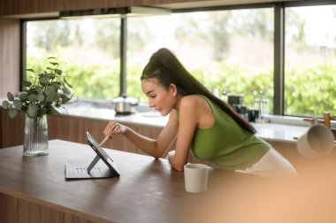 Genç Asyalı kadın evdeki mutfakta tabletle çalışıyor.