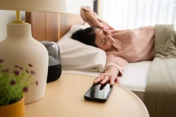 Sleeping asian woman turning off alarm on smartphone while being Waken up in the morning