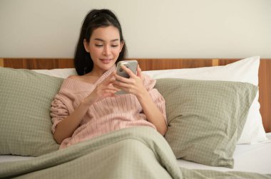 Genç Asyalı bir kadın yatakta uzanmış akıllı telefon kullanıyor.