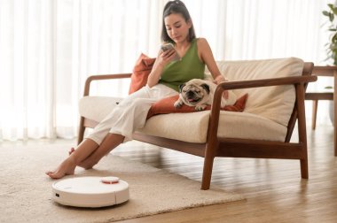 Robotik elektrikli süpürge çalışırken Asyalı genç bir kadın oturma odasında köpeğiyle dinleniyor.