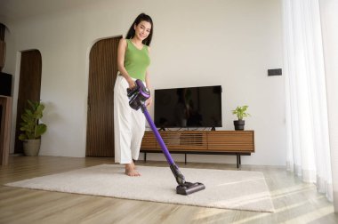 Genç ve mutlu bir kadın oturma odasının zeminini elektrikli süpürgeyle temizliyor.