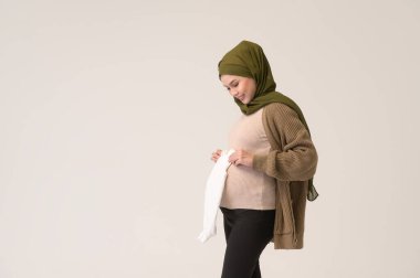 Beyaz arka plan stüdyosunun üzerinde bebek bezi tutan tesettürlü hamile Müslüman kadının portresi