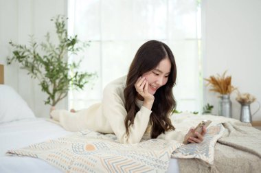Genç Asyalı kadın yatakta uzanmış akıllı telefon kullanıyor.