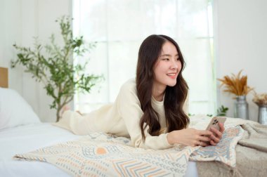 Genç Asyalı kadın yatakta uzanmış akıllı telefon kullanıyor.