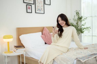 Genç Asyalı kadın yatakta uzanmış akıllı telefon kullanıyor.