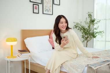 Genç Asyalı kadın yatakta uzanmış akıllı telefon kullanıyor.