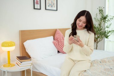 Genç Asyalı kadın yatakta uzanmış akıllı telefon kullanıyor.