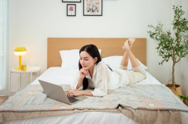Genç Asyalı kadın yatak odasında dizüstü bilgisayar kullanmaktan zevk alıyor.
