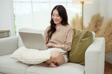 Genç Asyalı kadın oturma odasında dizüstü bilgisayar kullanmaktan hoşlanıyor.