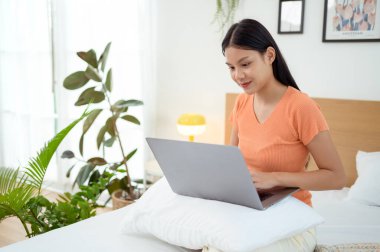 Genç Asyalı kadın yatak odasında dizüstü bilgisayar kullanmaktan zevk alıyor.