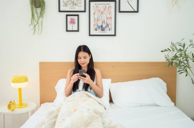 Genç Asyalı kadın yatakta uzanmış akıllı telefon kullanıyor.