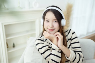 Kulaklık takan genç Asyalı kadın evdeki oturma odasında müzik dinliyor.