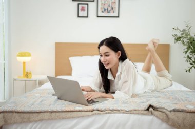 Genç Asyalı kadın yatak odasında dizüstü bilgisayar kullanmaktan zevk alıyor.