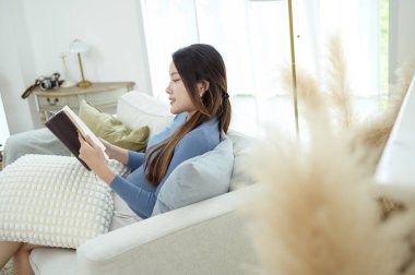 Genç Asyalı kadın oturma odasında kitap okumaktan zevk alıyor.