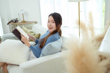 Genç Asyalı kadın oturma odasında kitap okumaktan zevk alıyor.