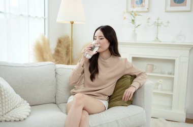 Genç Asyalı kadın evdeki oturma odasında su içiyor.