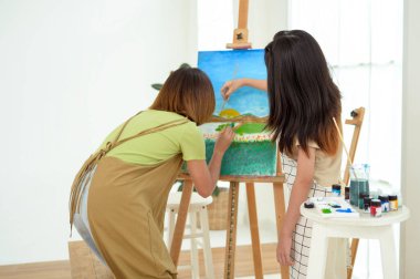 İki genç kadın, iyi aydınlatılmış bir sanat stüdyosunun sehpalarına manzara resmi çiziyor..