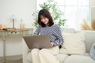 Huzurlu bir oturma odasında dizüstü bilgisayarıyla yazarken gülümseyen bir kadın..