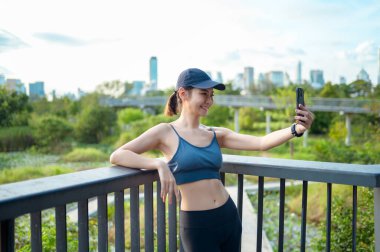 Aktif kıyafetli genç bir kadın şehirde güneşli bir günün tadını çıkarırken akıllı telefonunu kullanıyor..