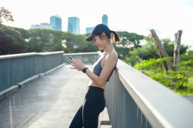 Aktif kıyafetli genç bir kadın şehirde güneşli bir günün tadını çıkarırken akıllı telefonunu kullanıyor..