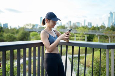 Aktif kıyafetli genç bir kadın şehirde güneşli bir günün tadını çıkarırken akıllı telefonunu kullanıyor..