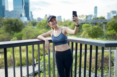 Aktif kıyafetli genç bir kadın şehirde güneşli bir günün tadını çıkarırken akıllı telefonunu kullanıyor..