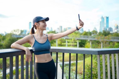 Aktif kıyafetli genç bir kadın şehirde güneşli bir günün tadını çıkarırken akıllı telefonunu kullanıyor..