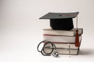 Siyahi bir mezuniyet şapkası steteskopun yanındaki istiflenmiş kitaplara dayanır, tıp eğitimini sembolize eder..