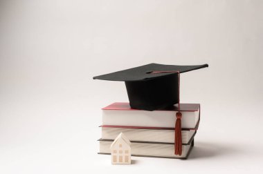 Siyah mezuniyet şapkası, eğitimi sembolize eden küçük bir ev figürünün yanındaki kitaplara dayanır..