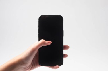 Beyaz bir arkaplana karşı aydınlatılmış, boş ekranlı siyah bir akıllı telefon tutan bir kişi var..