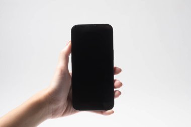 Beyaz bir arkaplana karşı aydınlatılmış, boş ekranlı siyah bir akıllı telefon tutan bir kişi var..