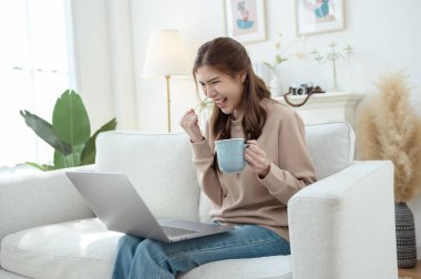 Genç bir kadın dizüstü bilgisayarına odaklanmış, elinde bir fincan kahveyle aydınlık bir oturma odasında.