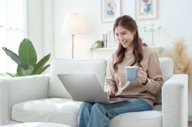 Genç bir kadın dizüstü bilgisayarına odaklanmış, elinde bir fincan kahveyle aydınlık bir oturma odasında.