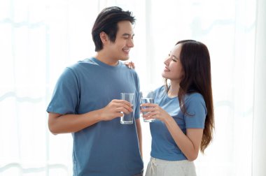 İki gülümseyen birey bir bardak su tutuyor, güneşli bir odada birbirlerine eşlik ederek eğleniyorlar..