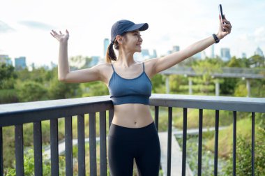 Aktif kıyafetli genç bir kadın şehirde güneşli bir günün tadını çıkarırken akıllı telefonunu kullanıyor..